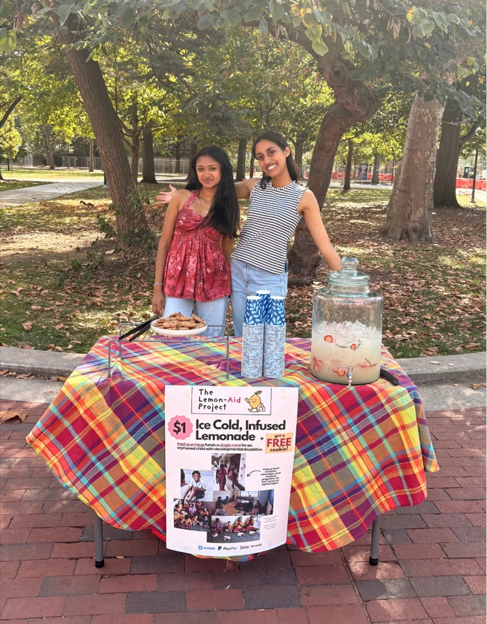 Ksheetisha and Yukta behind a Lemon-Aid stand on campus, with lemonade, cookies and a project poster
